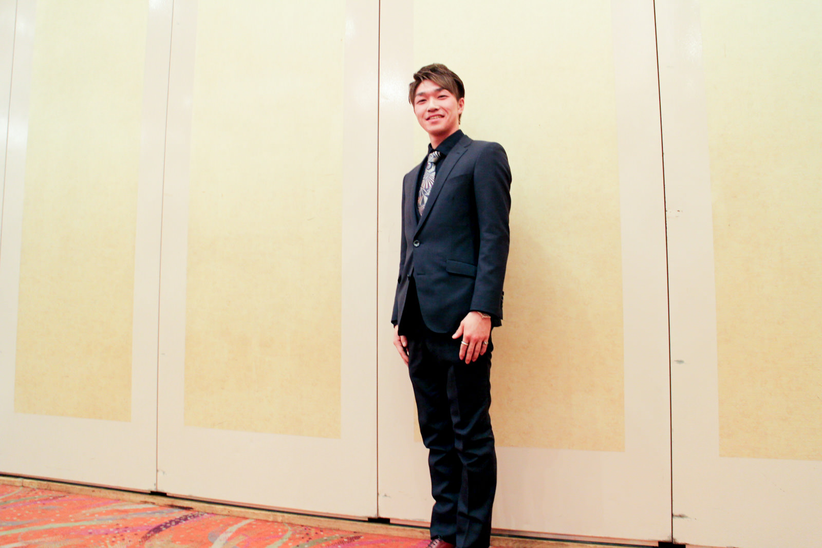 ベージュ色の壁の前で、黒いスーツに柄のネクタイを締めた新成人の男性が笑顔で左斜めに立っている写真