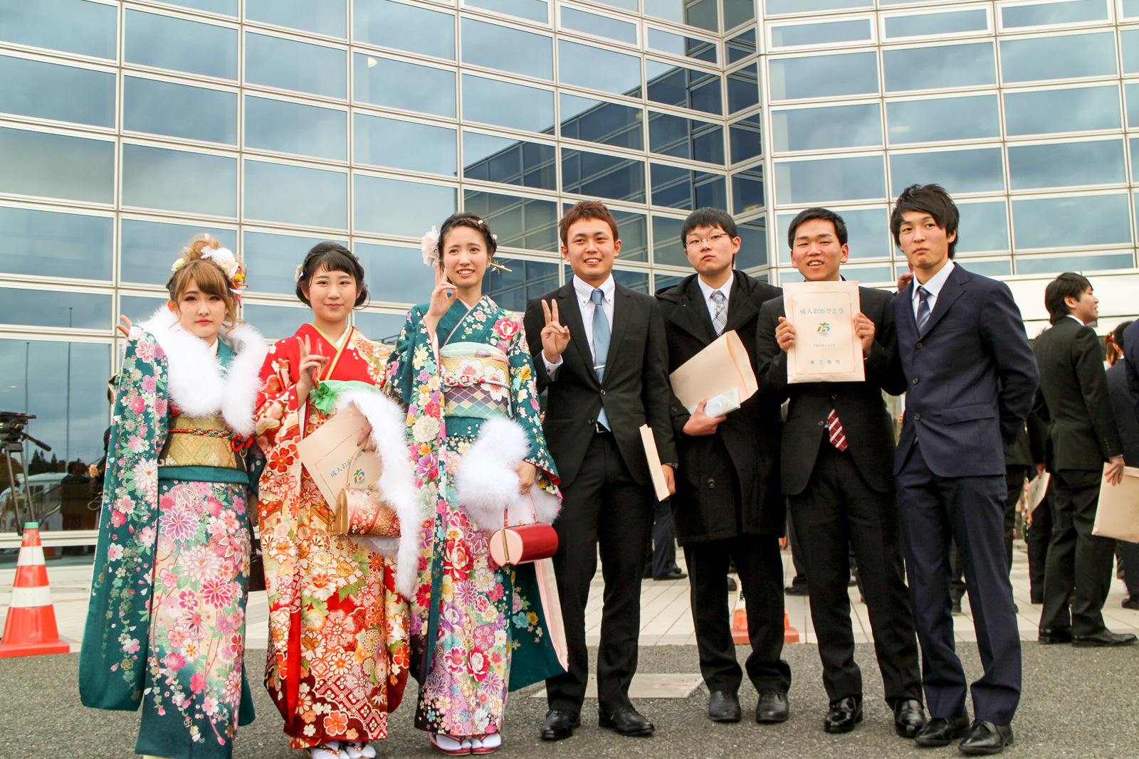 ガラス張りの建物の前で、振袖姿の女性3人とスーツ姿の男性4人が並び、封筒を手にしながらピースサインで微笑んでいる写真