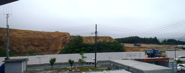 道路沿いの白いフェンスの中に土の山があり、右側の平地にはトラックなどの車両が見える写真