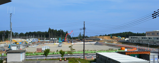 道路と黄緑色の低いフェンスで隔てられた建設現場の広い敷地に重機やトラックがあり、右側には仮設の建物が建っている様子を写した写真
