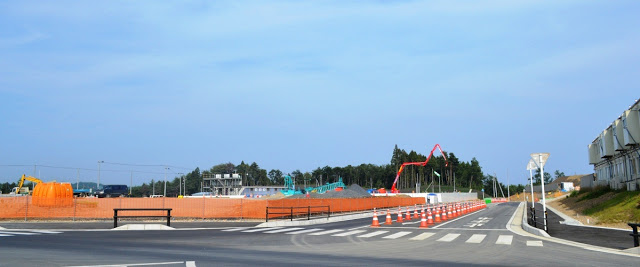 横断歩道がある道路の中央にカラーコーンが並んでおり、道路の左側のオレンジ色のフェンスで囲まれた建設現場の中に重機や建設資材が見える様子を写した写真