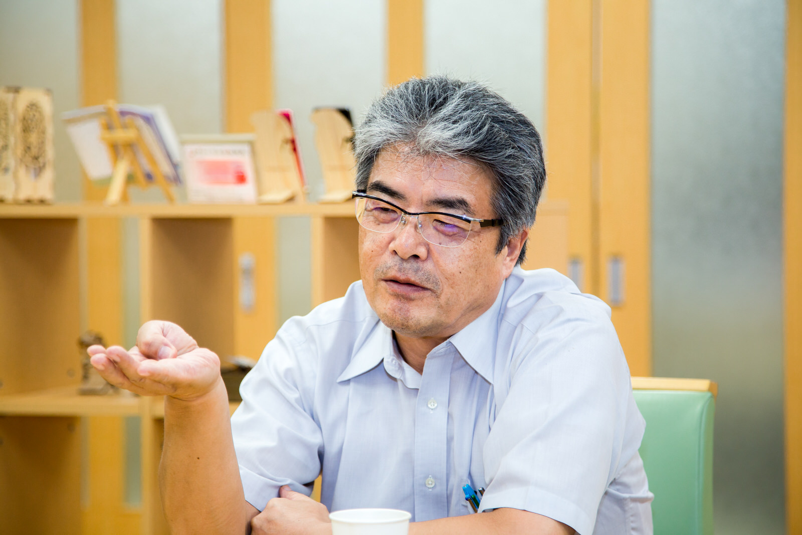 展示物が飾られた木の棚の前で、山内校長が、右手の親指を曲げて数を数えながら話している様子の写真