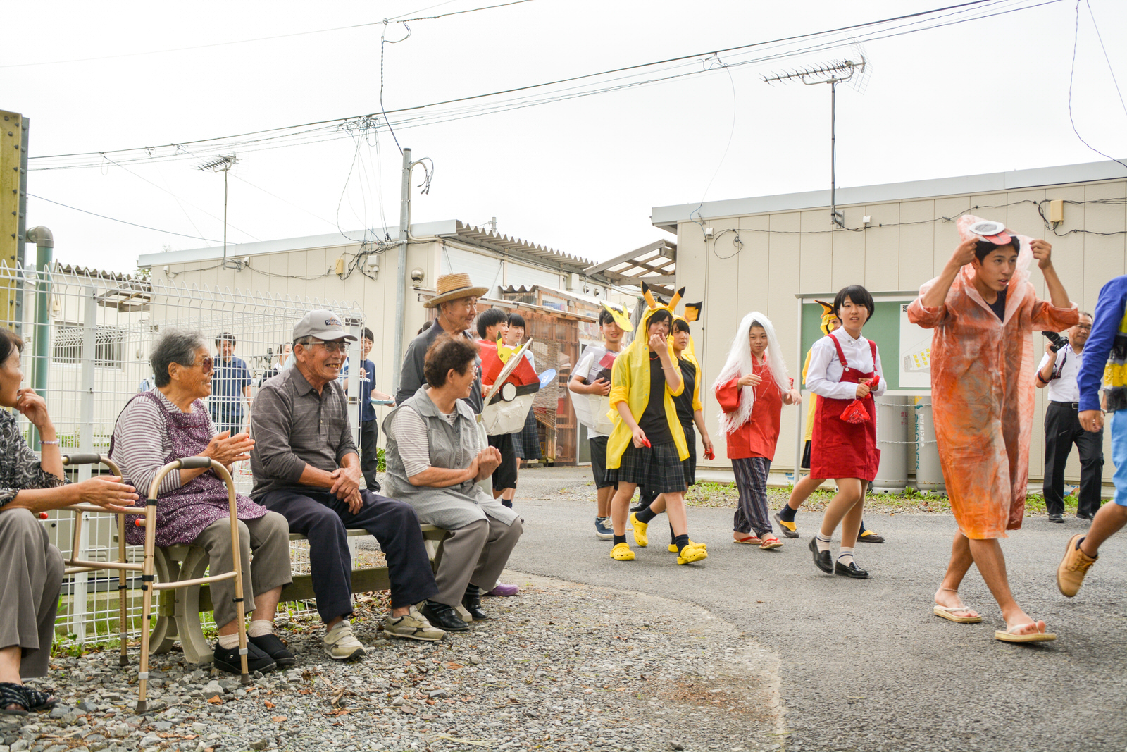 仮設住宅の敷地で、高齢者たちが椅子に座って見守る中、学生たちがアニメキャラクターの仮装をして楽しそうにパレードしている写真