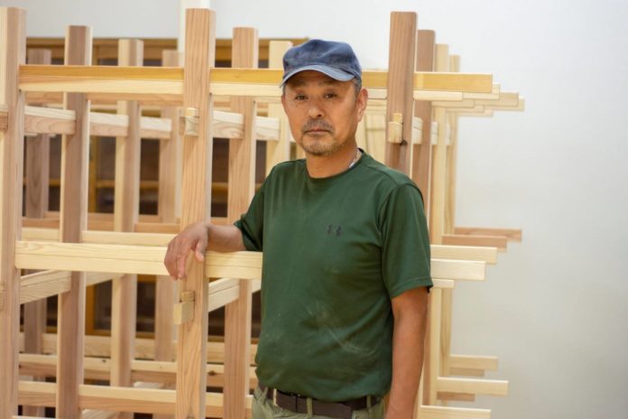 木材で組まれた棚のような構造物の前に立つ、緑色のTシャツと青い帽子をかぶった阿部さんの写真