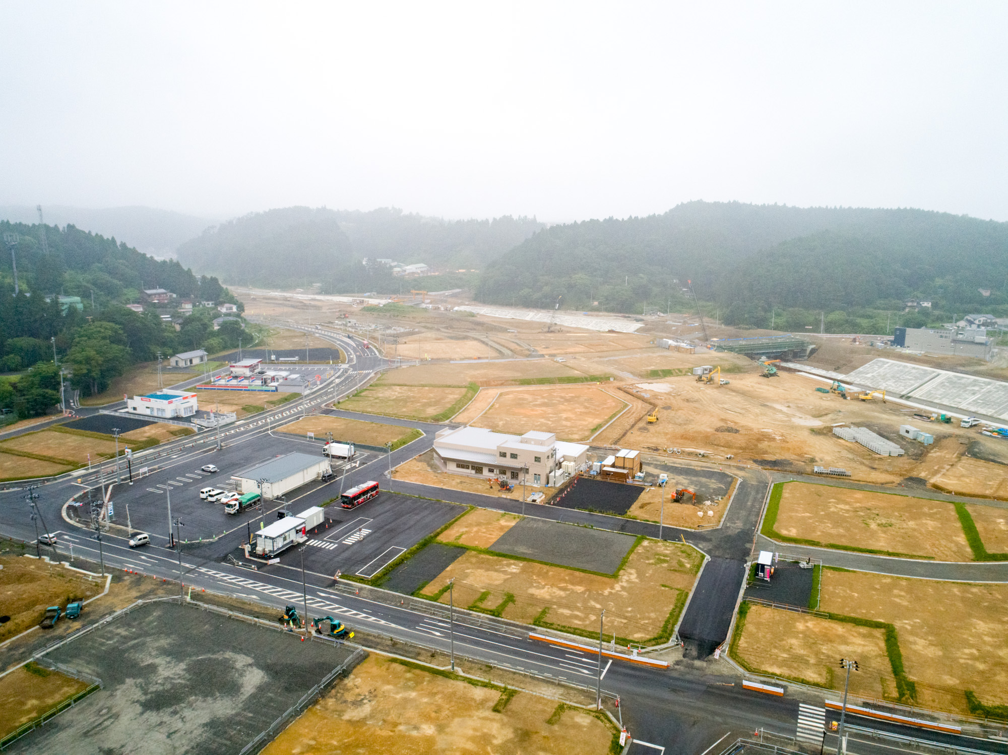 丘や木々に囲まれた地域で、複数の建物や道路、車両が点在する開発初期の工事現場を上空から捉え、区画された土地が今後の建設を予感させる、進行中の都市計画の様子を映した写真