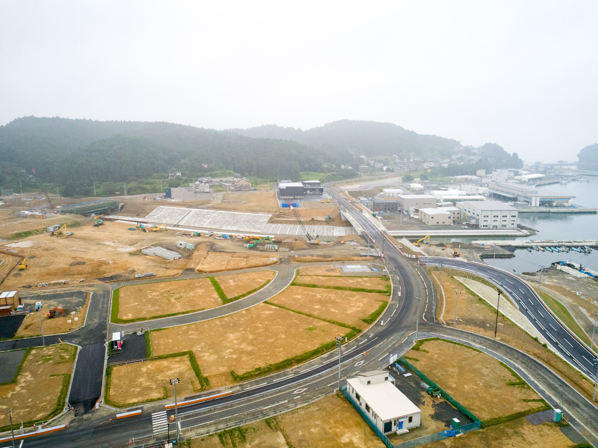 海岸近くの地域で、複数の道路が交差しながら整備・建設が進められており、区画された土地に建物や構造物が点在し、背景には木々に覆われた丘と沿岸の建物が広がる、インフラ開発が進行中の地域を上空から捉えた写真
