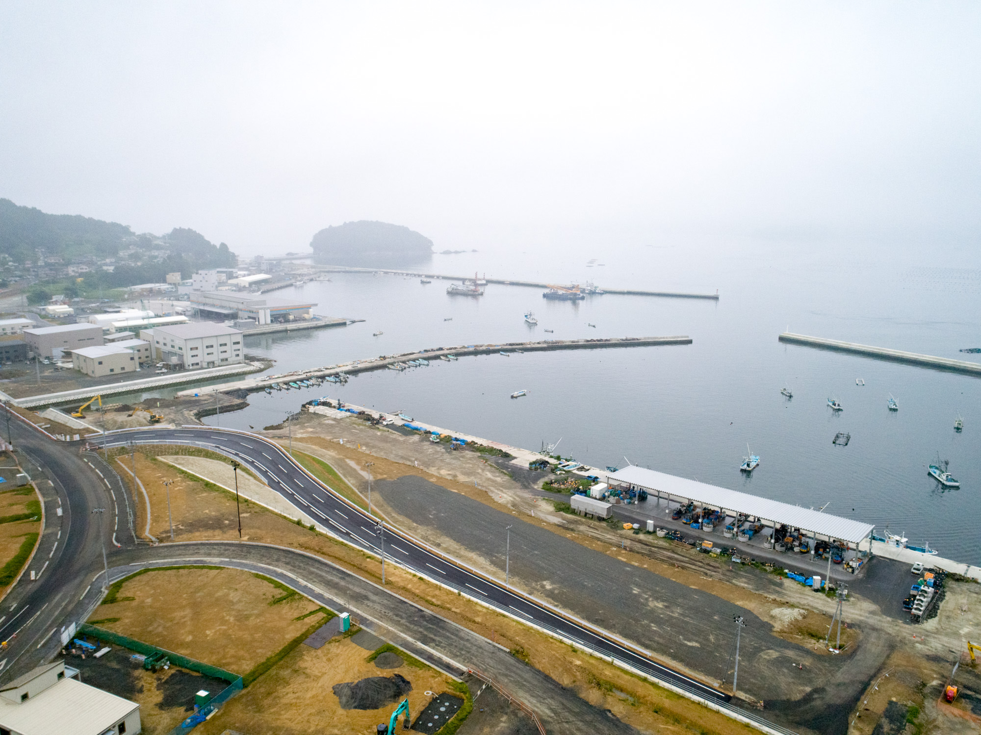 湾に沿って建物や構造物が立ち並び、湾内には複数の船が停泊しており、曲がりくねった道路や工事中のエリア、緑地が点在する海辺の地域を上空から捉えた、霧の中に小島も見える港町の写真