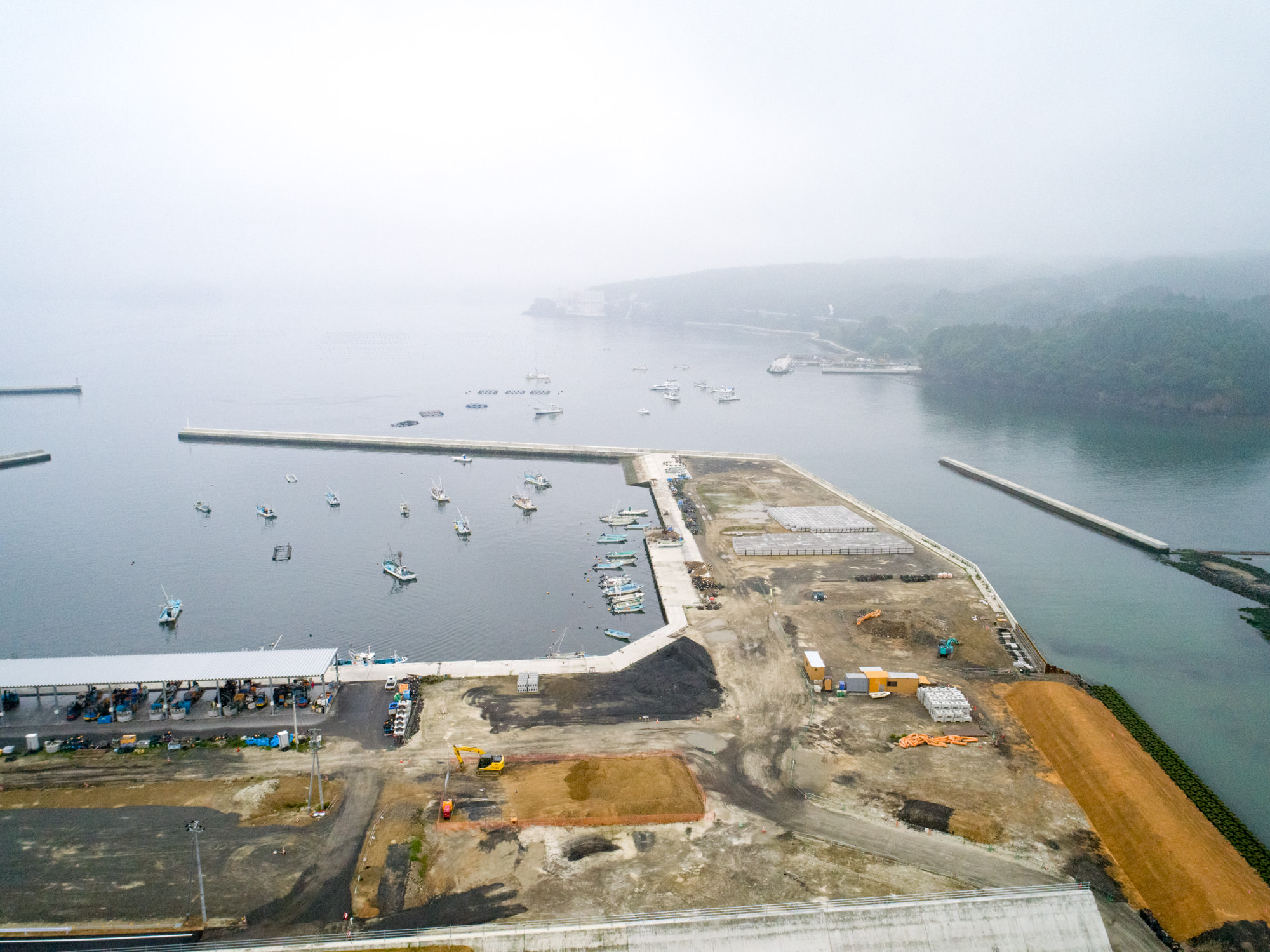霧がかった海辺の地域で、防波堤に囲まれた港に複数の船が停泊しており、隣接する陸地では建設資材や機材が並ぶ工事が進行中の様子を上空から捉えた、港湾開発の風景の写真