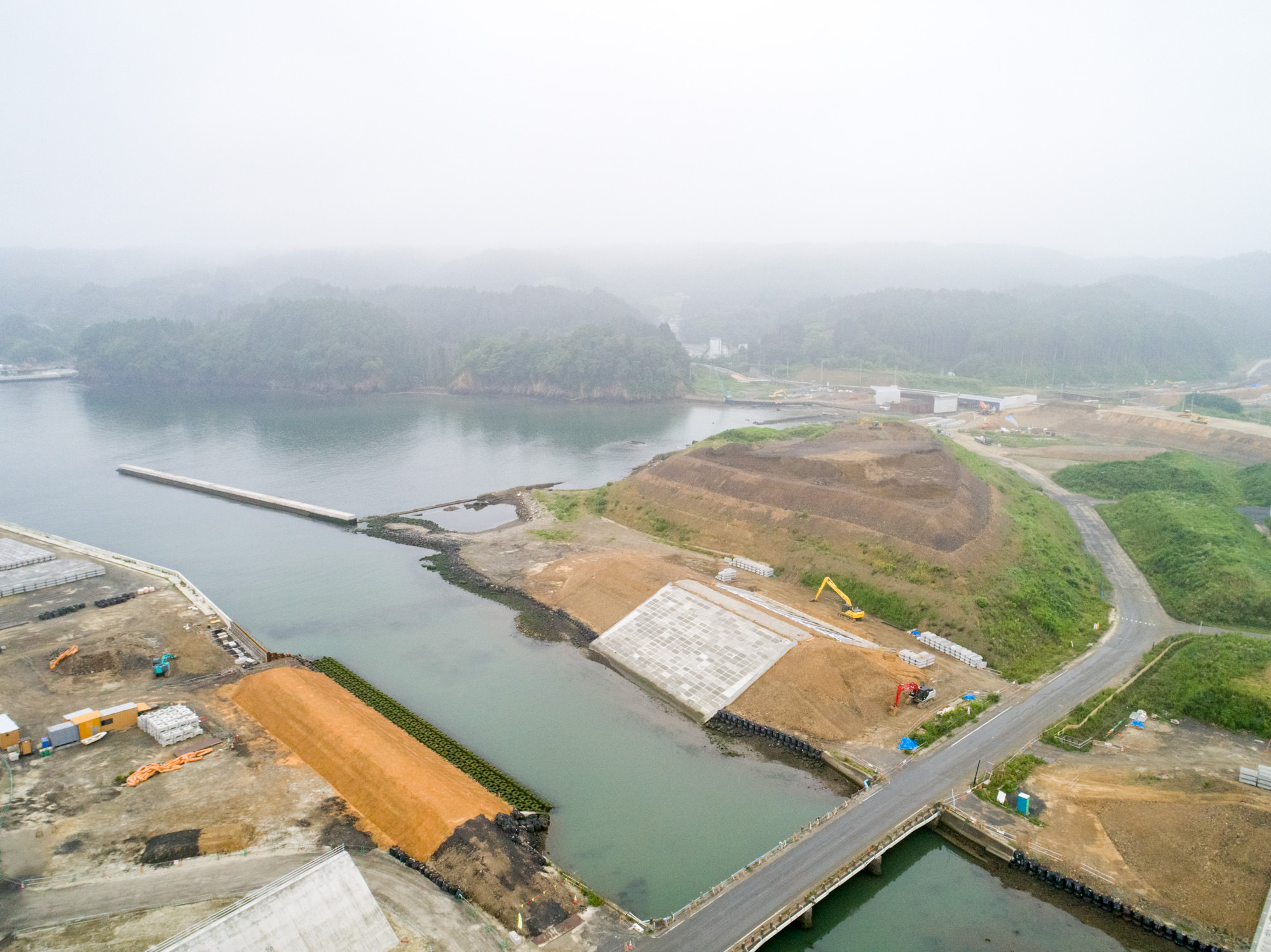 海沿いの開発地域で、むき出しの土やコンクリート構造物が点在し、道路が水辺にかかる小さな橋を通って伸びており、背景には緑の丘と建設機材が見える、水辺のインフラ整備が進む様子を上空から捉えた写真