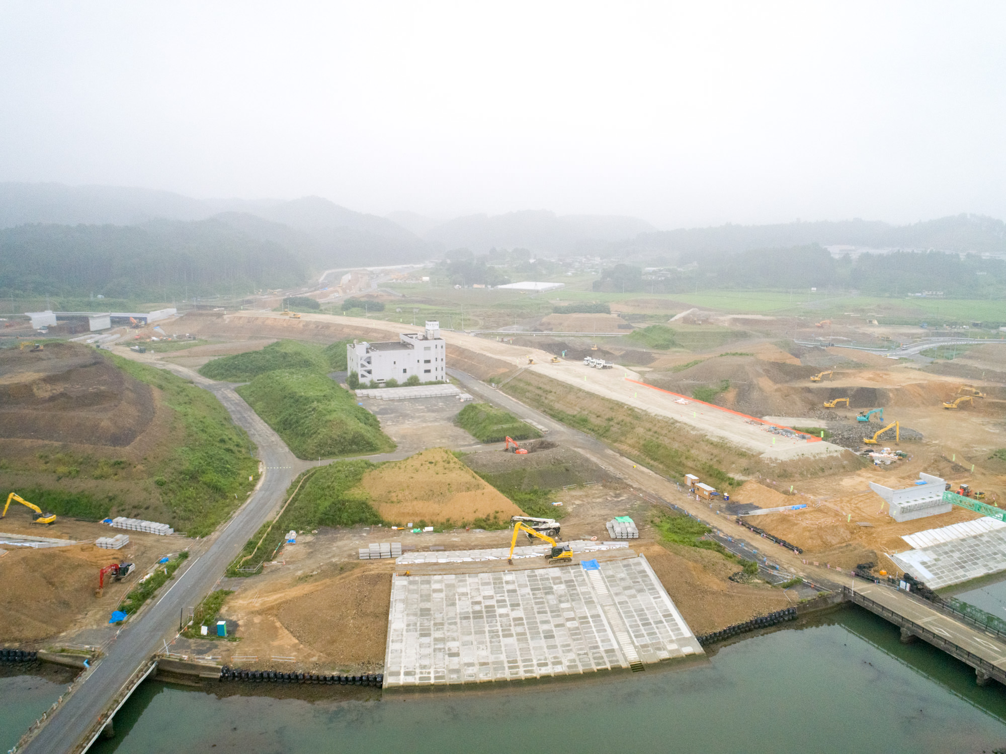 中央に大きな建物があり、周囲に道路や通路が広がる工事現場で、複数の重機や建設車両が稼働しており、背景には緑の丘が広がり、画面下には水辺も見える地域で進む水辺開発の様子を上空から捉えた写真