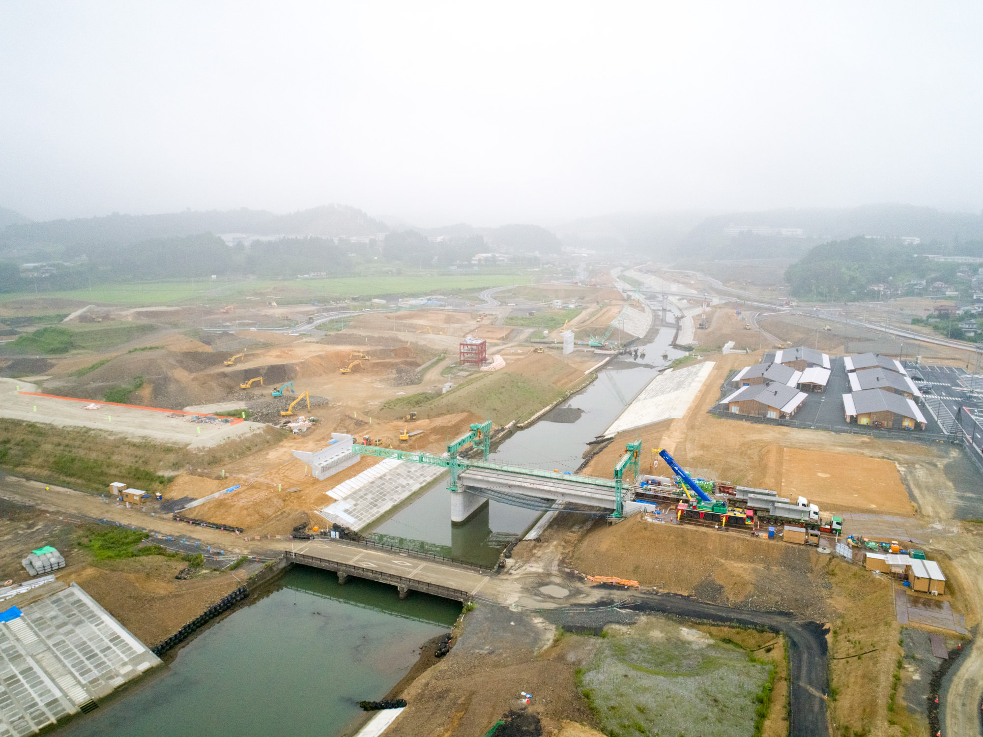 川が流れる広大な工事現場で、複数の橋が建設中であり、重機や建物が点在し、周囲を丘に囲まれた地域でインフラ整備が進む様子を上空から捉えた、開発中の風景の写真