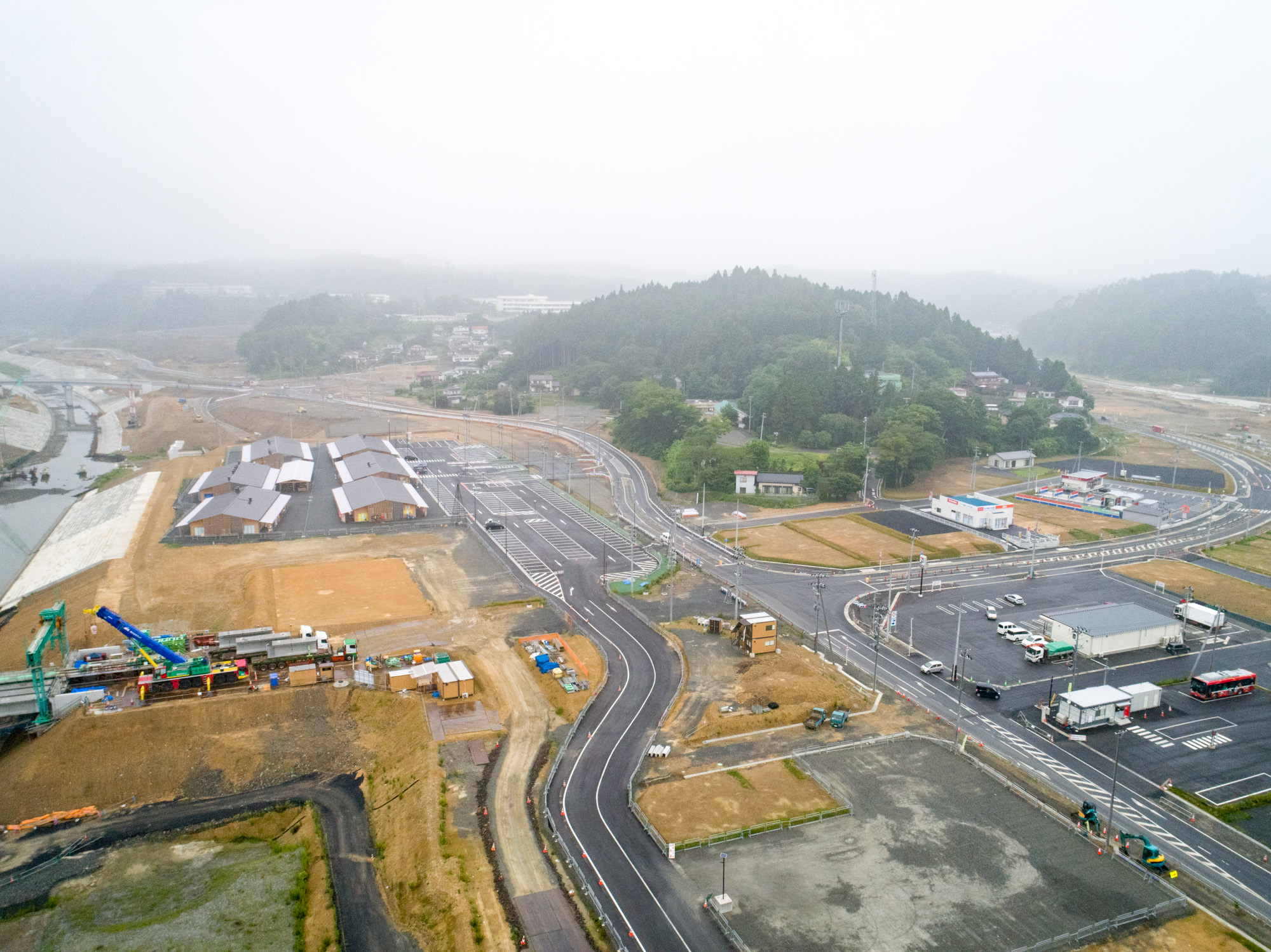 霧がかった空の下、木々に覆われた丘を背景に、舗装された新しい道路や建設中の建物、工事機材が点在し、整地された土地と自然が混在する地域を上空から捉えた工事現場の写真