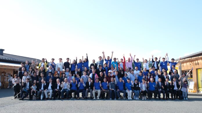 晴天の空の下、多くの人々が屋外で集合し、笑顔で手を挙げたポーズの記念写真