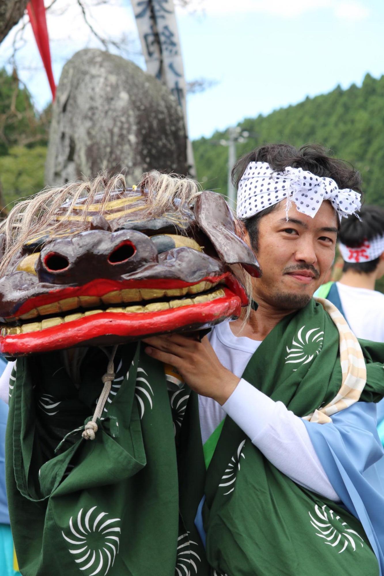 屋外の祭りの場で、緑色の獅子舞の装束をまとい白い鉢巻を締めた男性が、大きな赤い口と金色の歯を持つ獅子頭を肩に担ぎ、晴れやかな表情でこちらを見ている写真