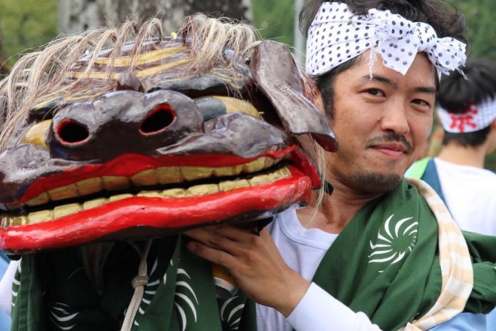 屋外の祭りの場で、緑色の法被をまとい白い鉢巻を締めた男性が、大きな赤い口と金色の歯を持つ獅子頭を肩に担ぎ、晴れやかな表情でこちらを見ている写真