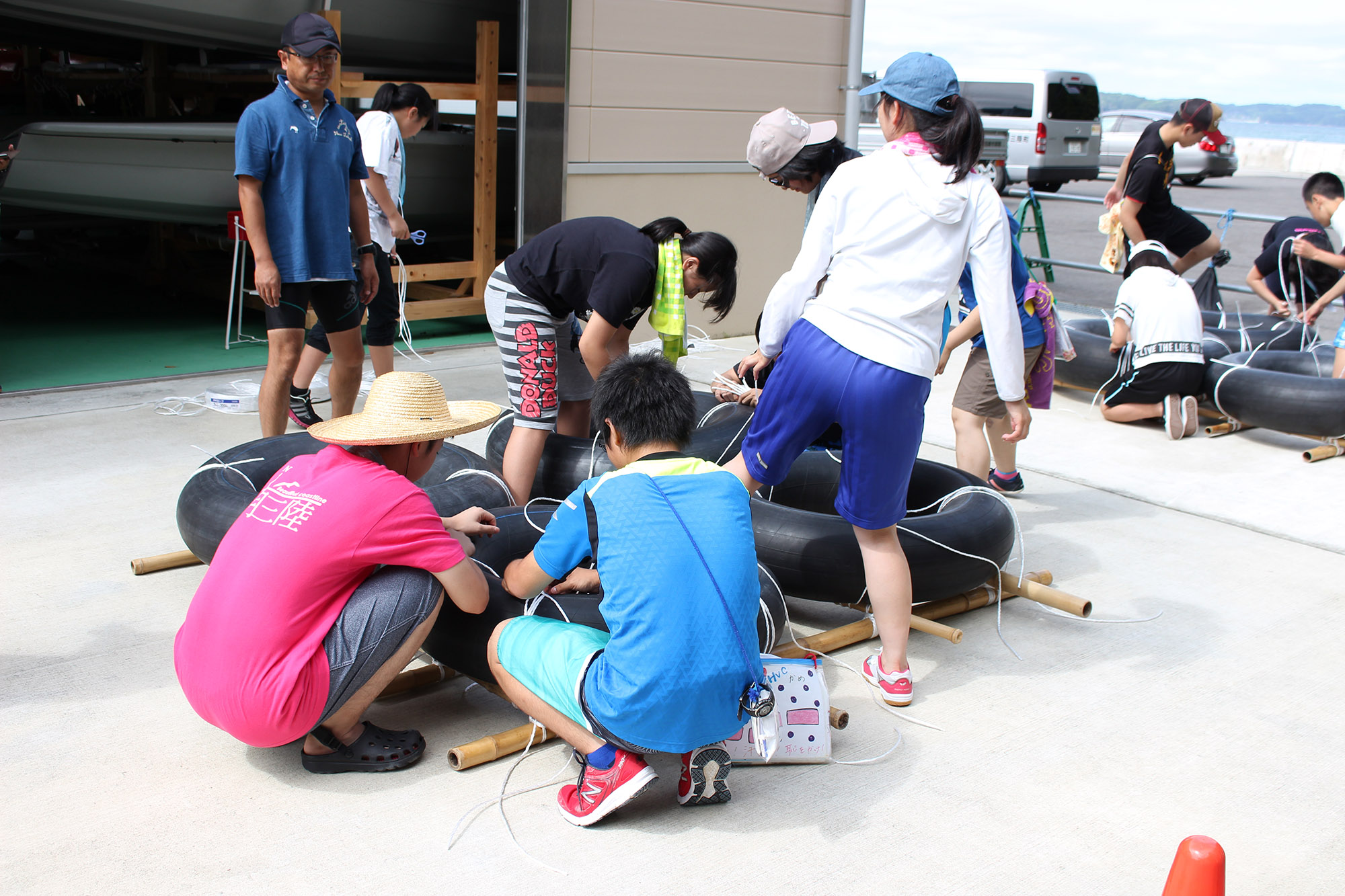 男性が見守る中、5名ほどのジュニアリーダーたちが、竹とタイヤチューブとロープを使って、協力しながらいかだ作りを行っている写真