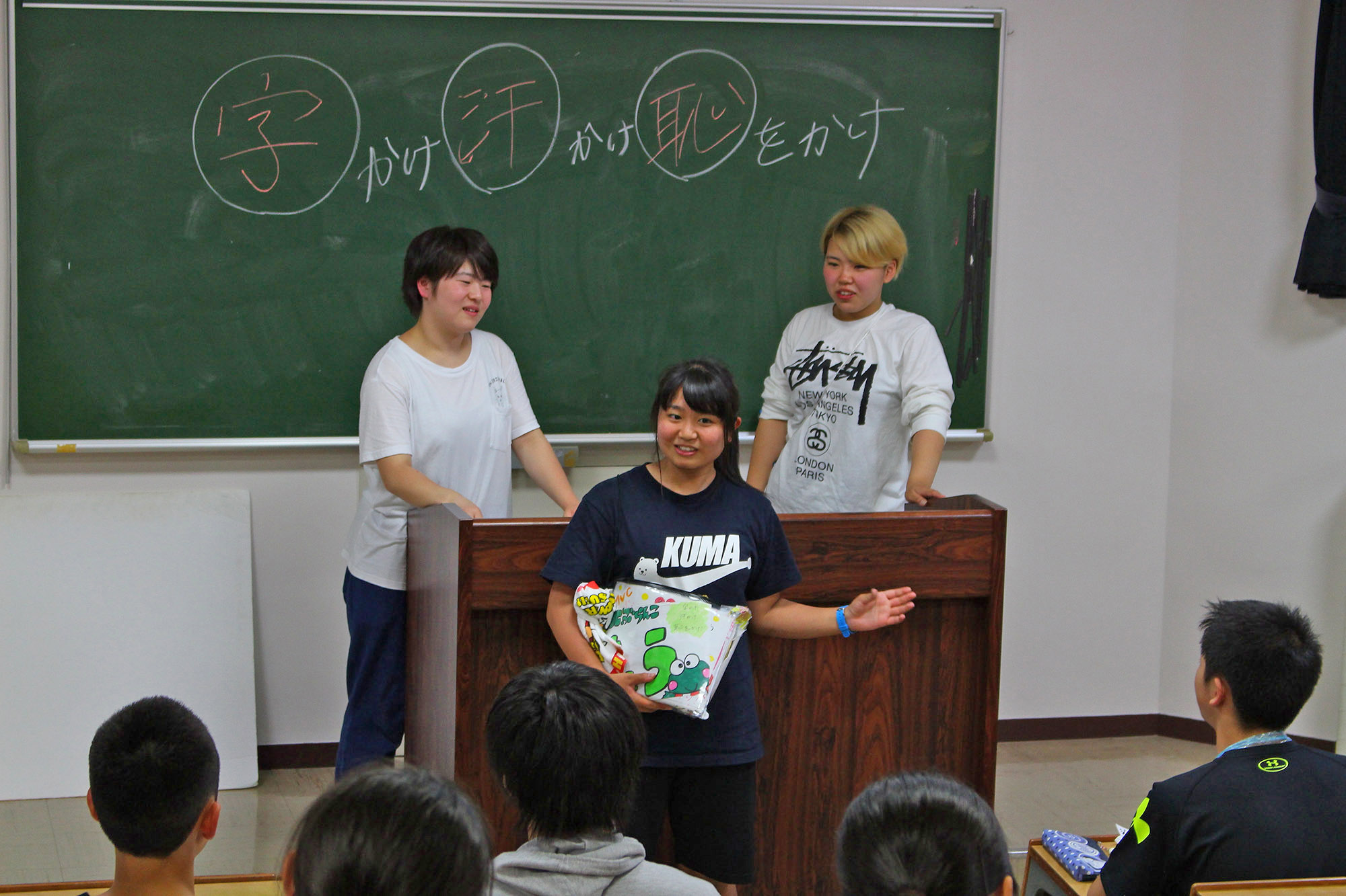 教室の黒板の前で「字を書け」「汗をかけ」「恥をかけ」と書かれた言葉を背に三人の若者が発表しており、他の学生たちは座って発表を見ている様子の写真