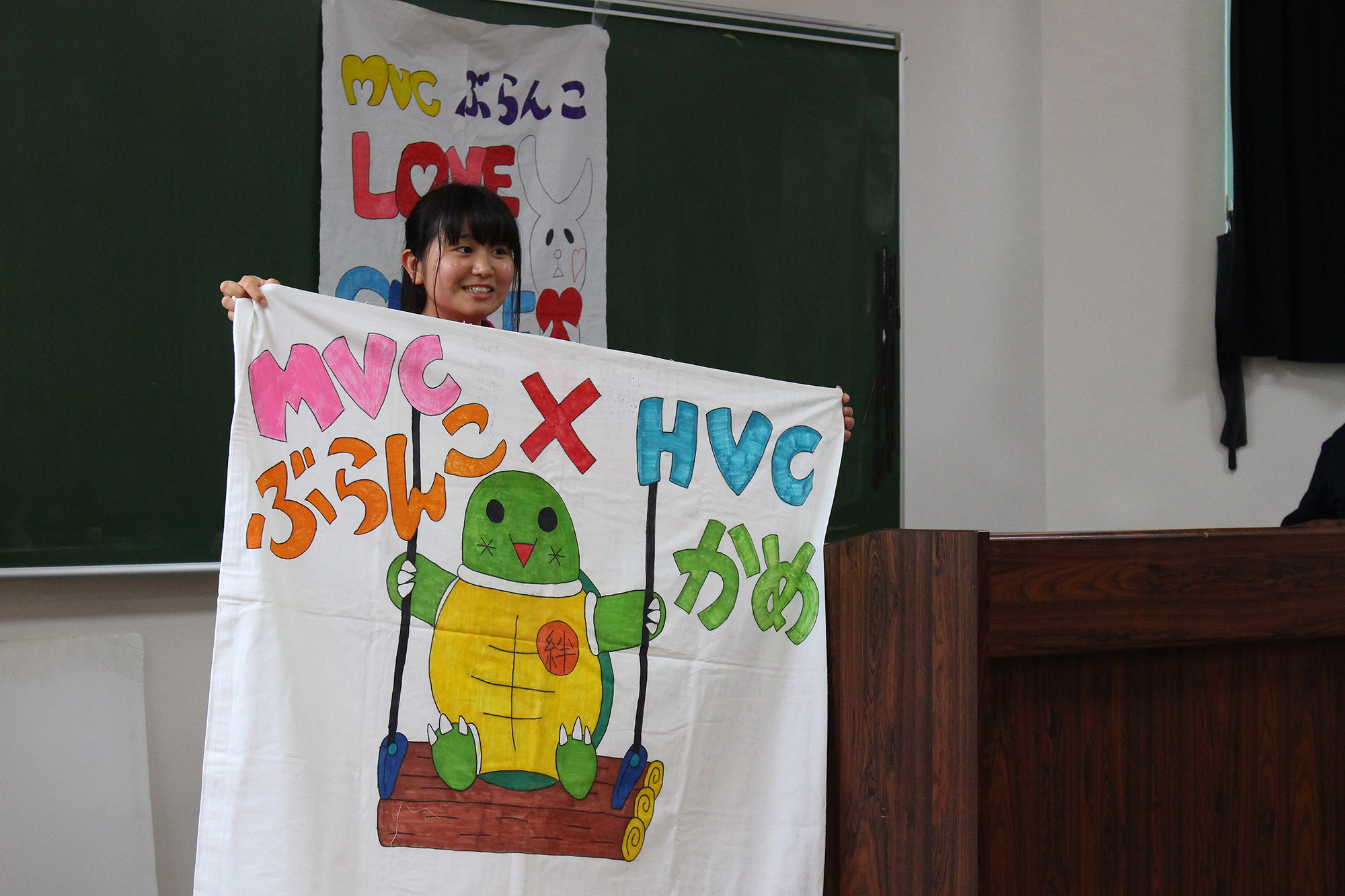 女子生徒が「MVC ぶらんこ × HVC かめ」と書かれ、ブランコに乗っているカメが描かれた旗を手に持ち立っている写真