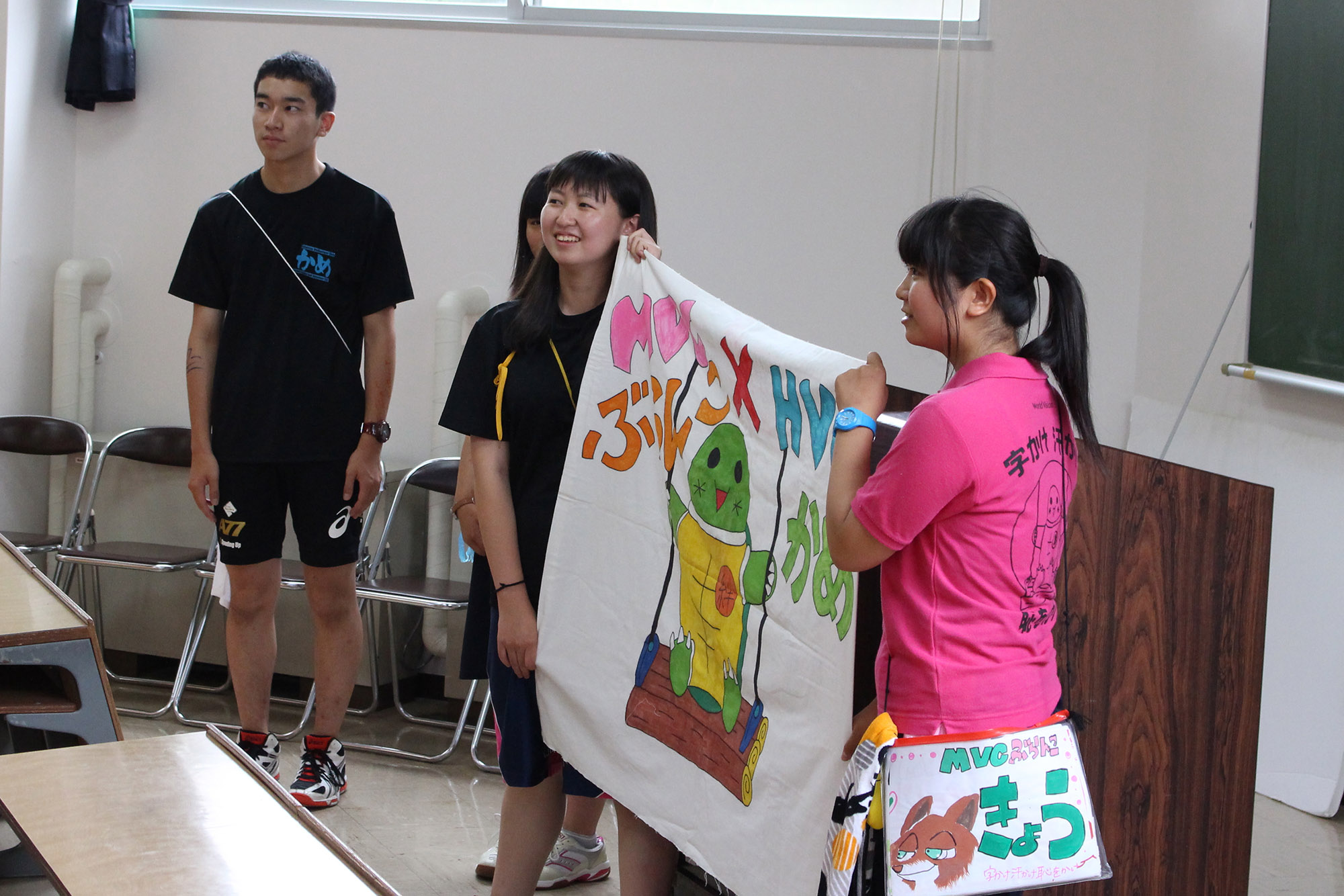 室内の前方で、ブランコに乗ったキャラクターと「ぶらんこ」などの文字が描かれているカラフルな手描きの旗を広げて笑顔で立っているジュニアリーダーの女子学生2人を写した写真