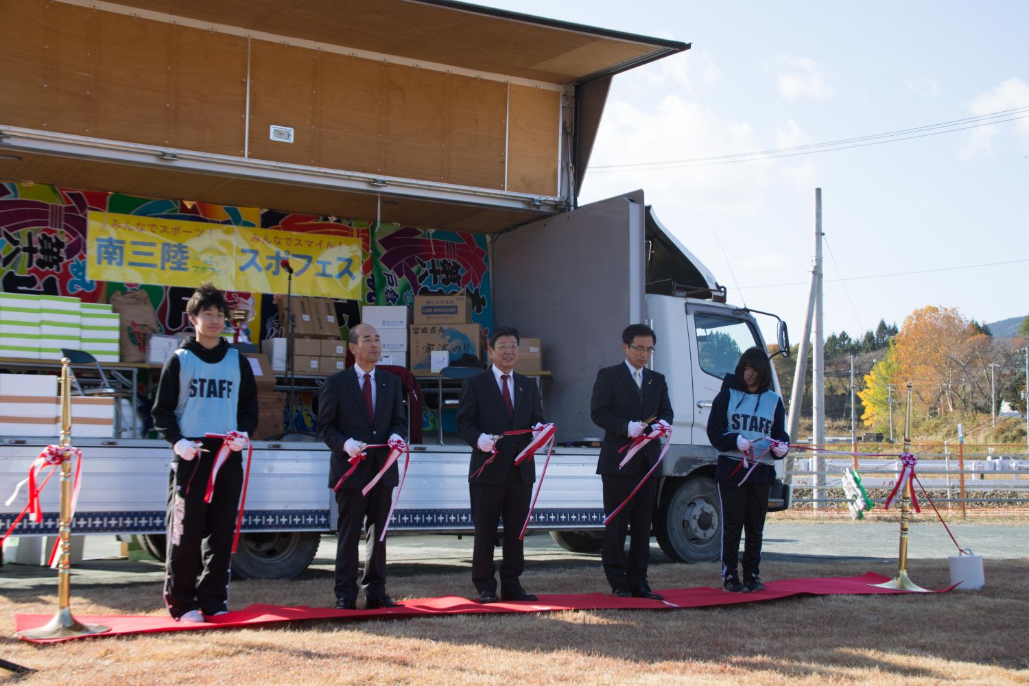 「南三陸スポフェス」と書かれた黄色の横断幕が掲げられたトラックの荷台に段ボールや箱が積まれ、水色のビブスを着たスタッフと3人のスーツ姿の男性がレッドカーペットの上に間隔をあけて立ちテープカットが行われている様子の写真