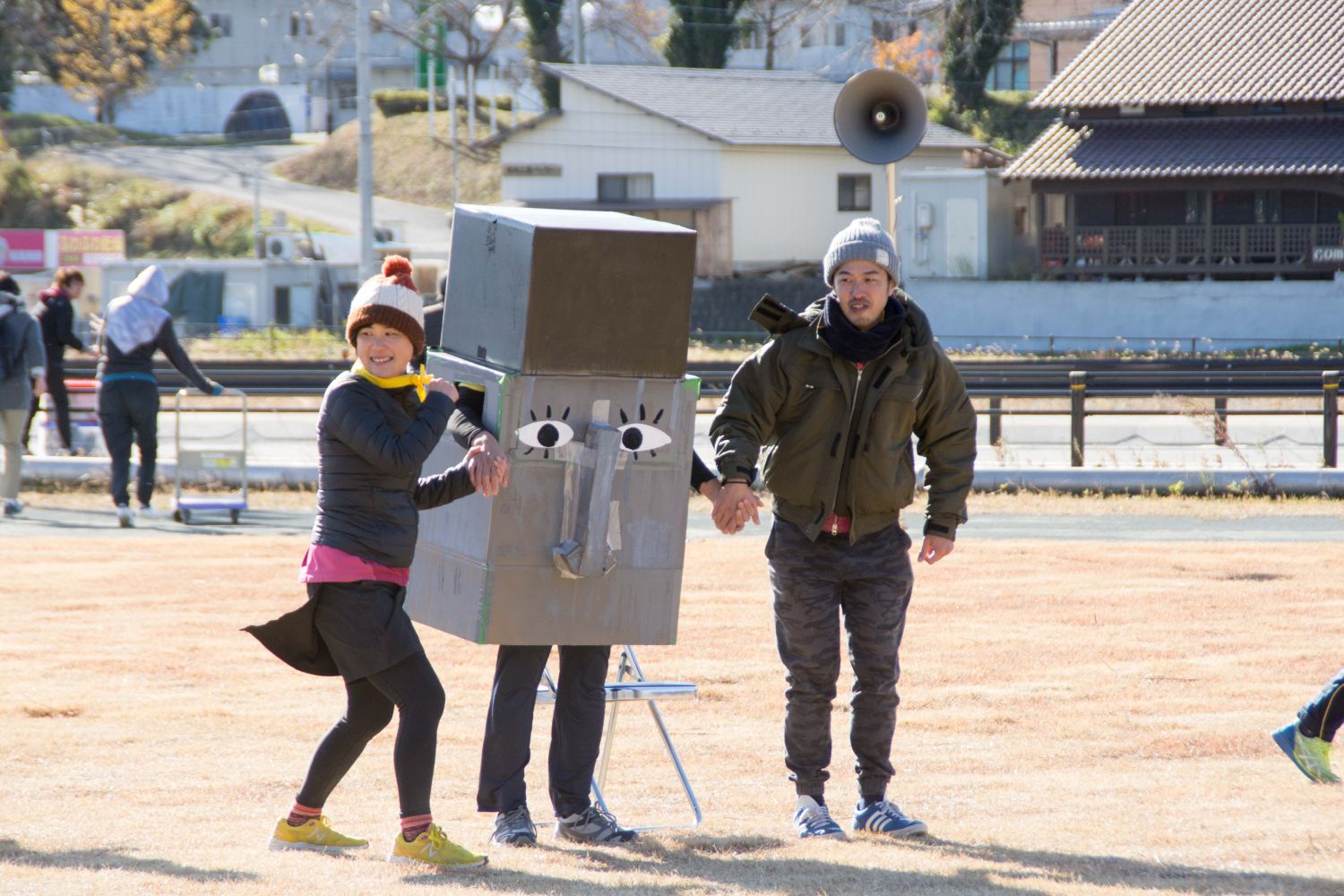 長方形と四角形の箱で作られたモアイの被り物を着たメンバーを中央に、2人と手を握って一緒に走りリレーを行っている様子の写真