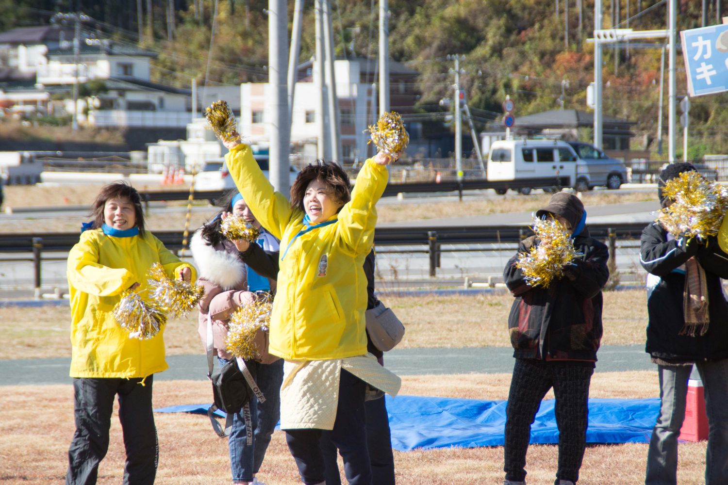 黄色のジャケットを着た女性の参加者が、金色と銀色のボンボンを両手で持ち応援している様子の写真