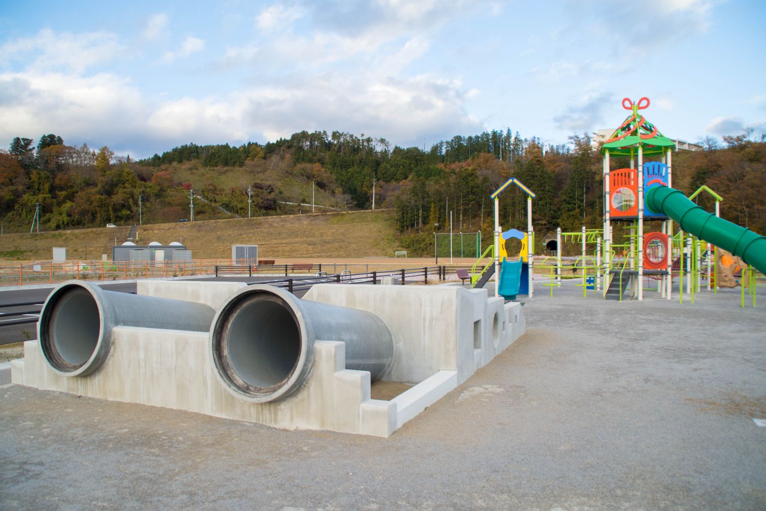 コンクリート造りの土台に2つの土管の様な物が並列し、奥にカラフルな色の複合遊具が設置された公園の写真