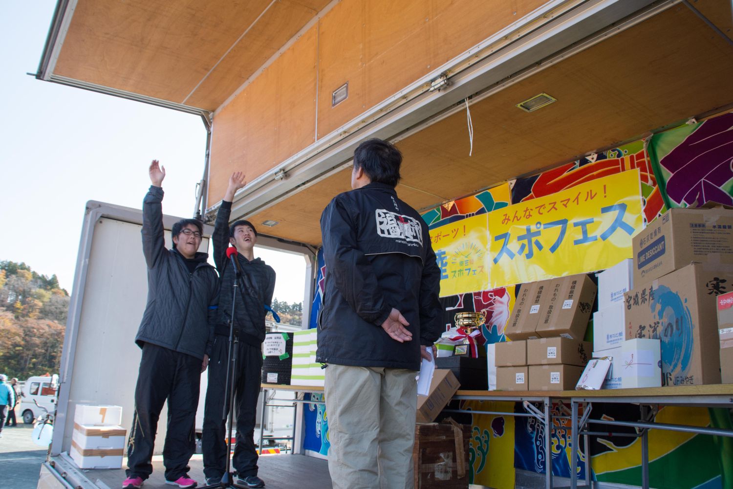 トラックの荷台に設置されたスタンドマイクの前で、黒色のスポーツウエアを着た2人の男性が右手を上に上げ選手宣誓を行っている様子の写真