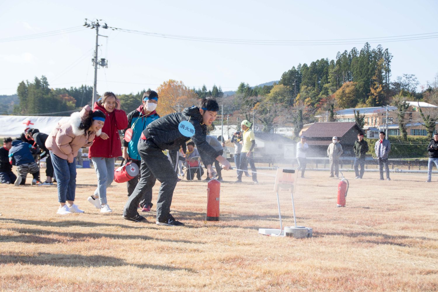 3人のメンバーが見ているなか、消火班と書かれた札を付けた男性が的に向かって消火器のノズルを向け初期消火を行っている様子の写真