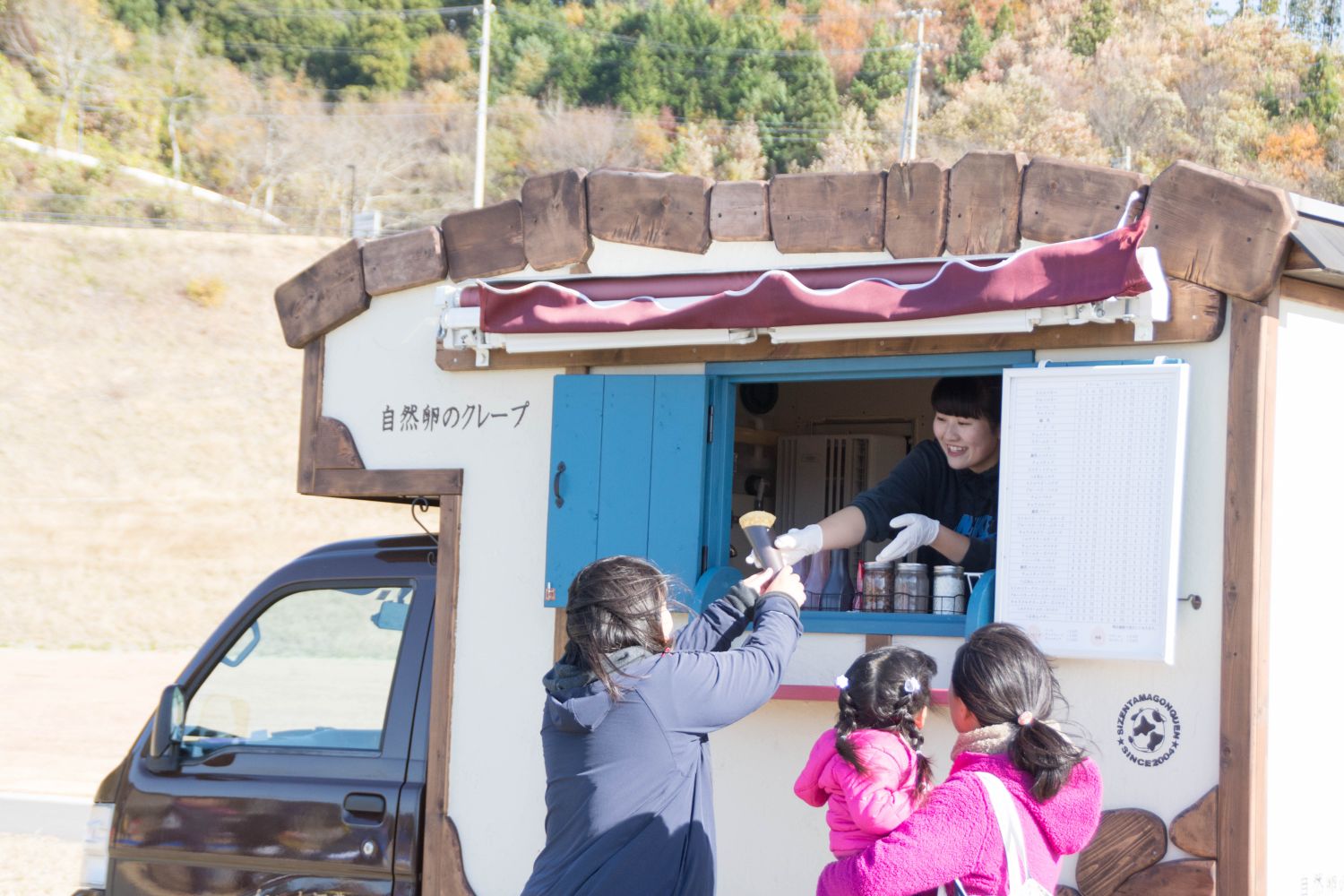 茶色のボディに白色をベースにした「自然卵のクレープ」と書かれたキッチンカーの窓から、出来上がったクレープを受け取る女性客の写真