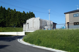 緩やかな坂道の道路沿いに草が生い茂り、左側の家は工事用シートで覆われている住宅地の写真