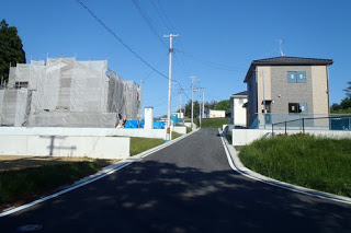 緩やかな坂道の両側に家が建ち並び、奥に電柱や青空が見える新興住宅地の写真