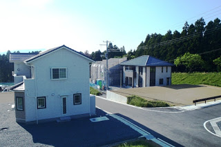 道路の両側に白い家やグレーの家が建っていて、周囲に緑と空き地が広がる住宅街の写真