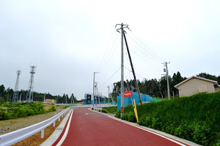赤く舗装された歩道の横に電柱と草の斜面があり、奥に青いネットの建設現場が見える写真