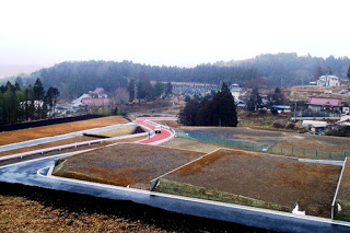 区画された宅地が手前に広がり、奥には赤い舗装の道路と山あいの集落が見える造成地の写真