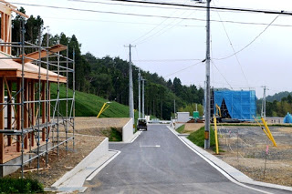 左側に木造の建設中の家があり、舗装道路が奥の青いネットの建物まで続く分譲地の写真