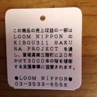 「この商品の売上収益の一部はLOOM NIPPONのKIBOU311 SAKURA PROJECTを通し、宮城県南三陸町に20年かけて3000本の桜を植樹する支援事業に協賛されます LOOM NIPPON 03-3533-6556」と書かれたタグが木の机に置かれている写真
