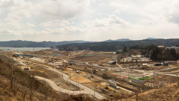 曲がりくねった道路があり、その周囲には複数の建設機械や車両が配置されいくつかの建物や構造物が点在し、さらに奥には山々と海が広がっている写真