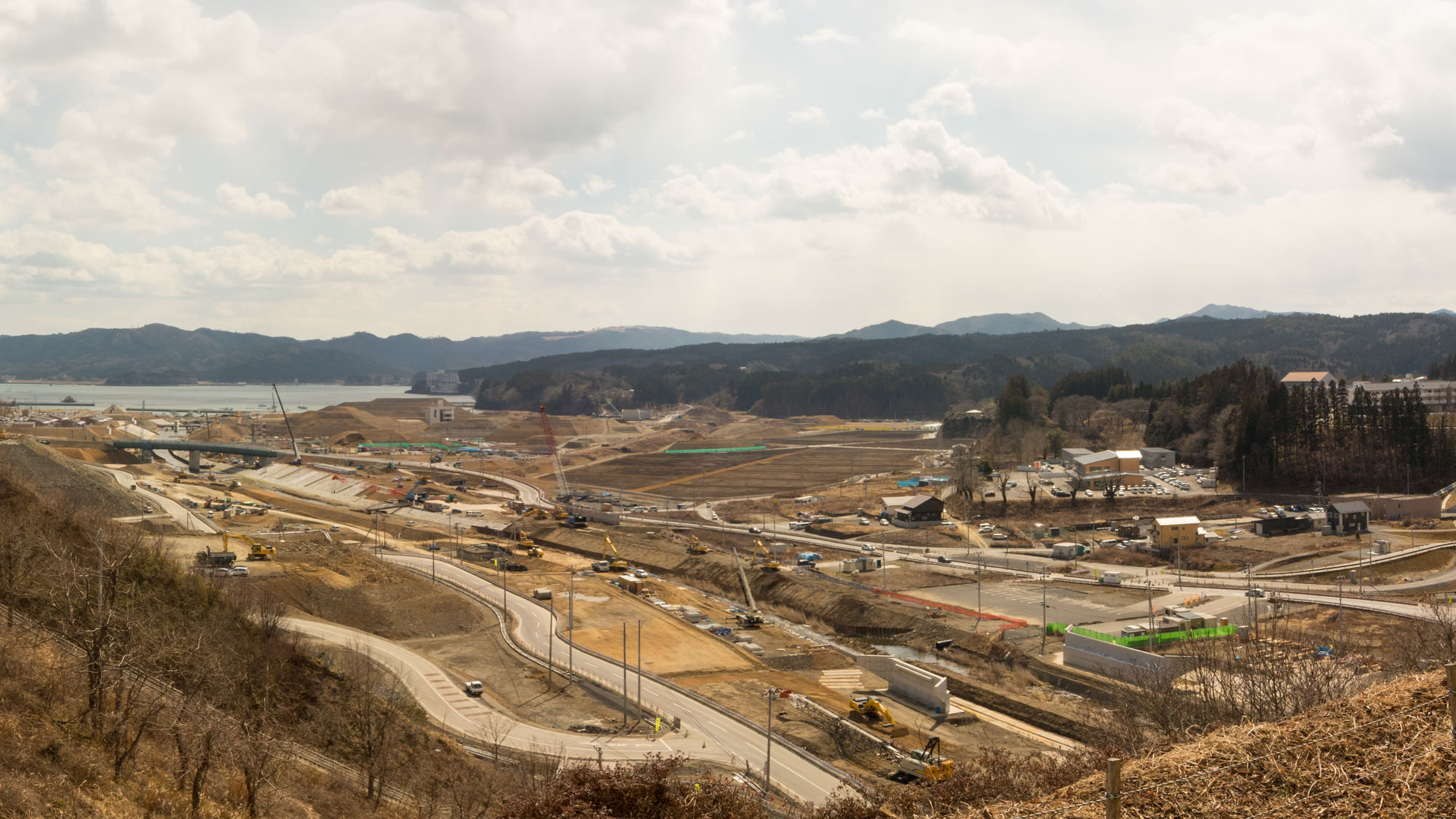 曲がりくねった道路があり、その周囲には複数の建設機械や車両が配置されいくつかの建物や構造物が点在し、さらに奥には山々と海が広がっている写真