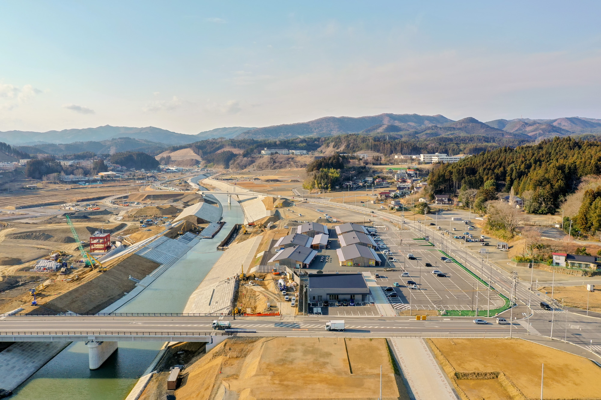 川に架かる橋を手前に、右側には建物や駐車場が広がり、背景には森林と丘陵が見える河川沿いの工事現場を上空から捉えた地域開発の写真