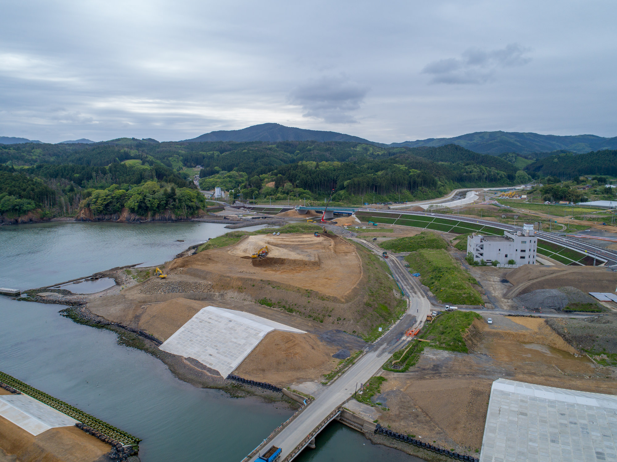 川辺に隣接した建設現場を上空から捉えた写真で、複数の大きな土の山と建設機材が見られ、水上には橋が架かり、その先に工事区域が広がり、背景には緑豊かな丘や山々が曇り空の下に広がる風景の写真