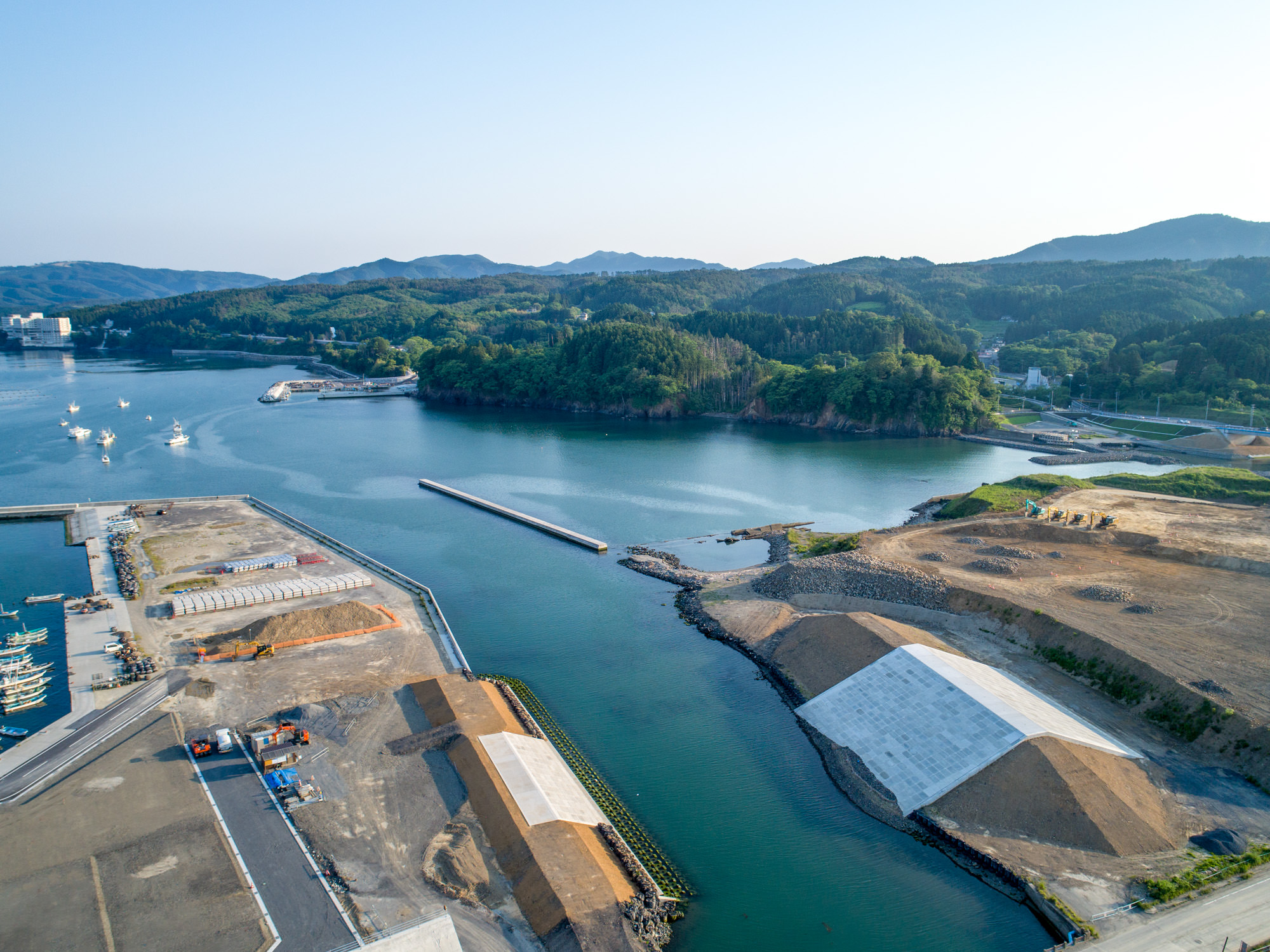 海に囲まれた地域を上空から撮影したもので、左側の水面には複数の船が浮かび、両岸には白いシートで覆われた盛土や建設資材・機材が点在し、背景には緑の丘と森林、青空が広がる晴天の写真