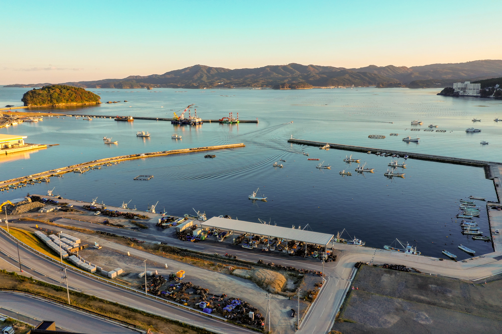 空から見下ろした海沿いの地域の風景で、港には複数の船や船舶が停泊していたり航行していたりし、港を囲む防波堤や海岸沿いの建物群が見え、背景には山々が広がり、空は澄んでいて朝焼けまたは夕焼けの暖かい光に包まれている写真