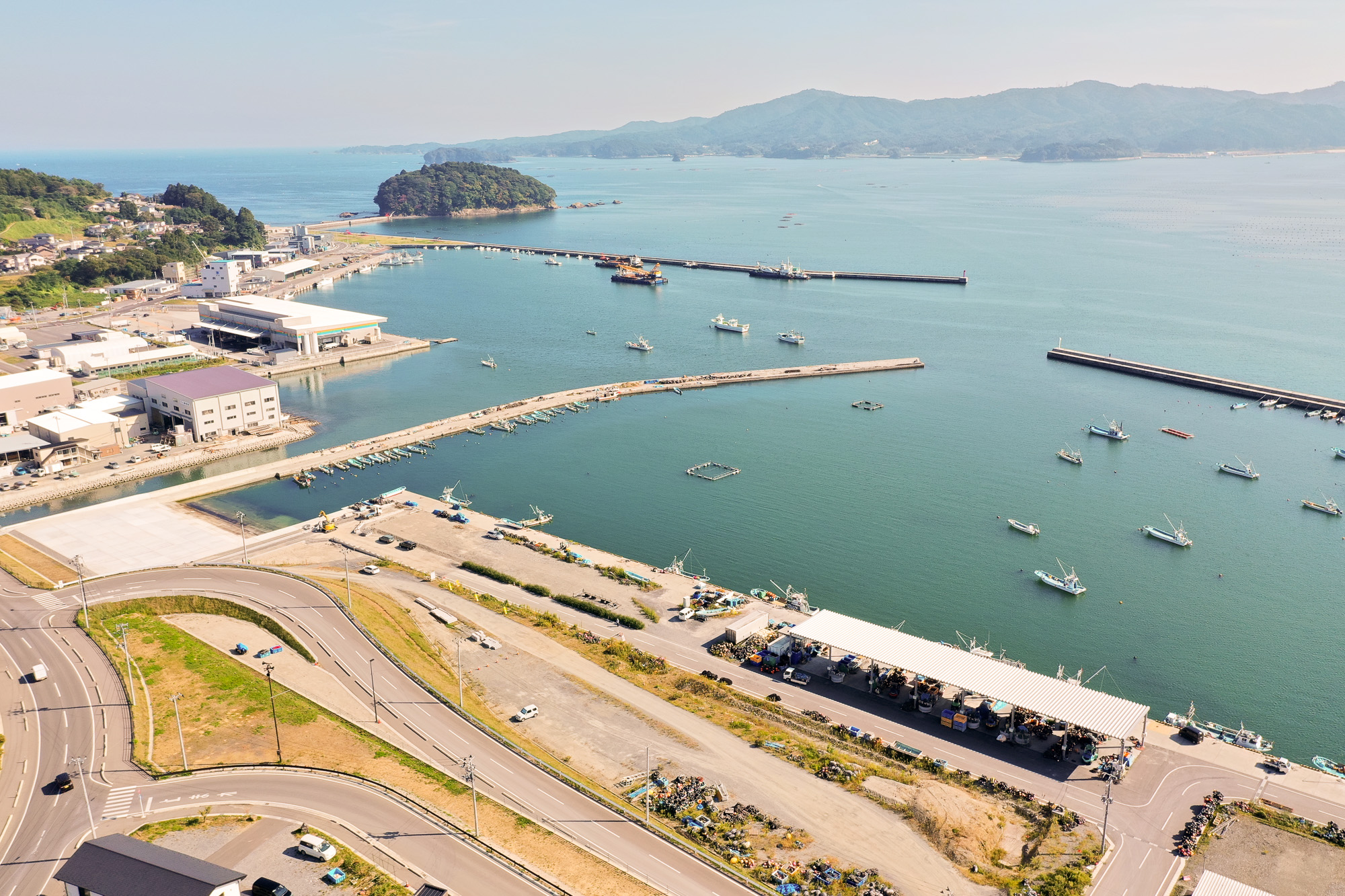 手前にカーブした道路と漁港施設があり、多数の漁船が停泊している様子を高台から見下ろした港の写真