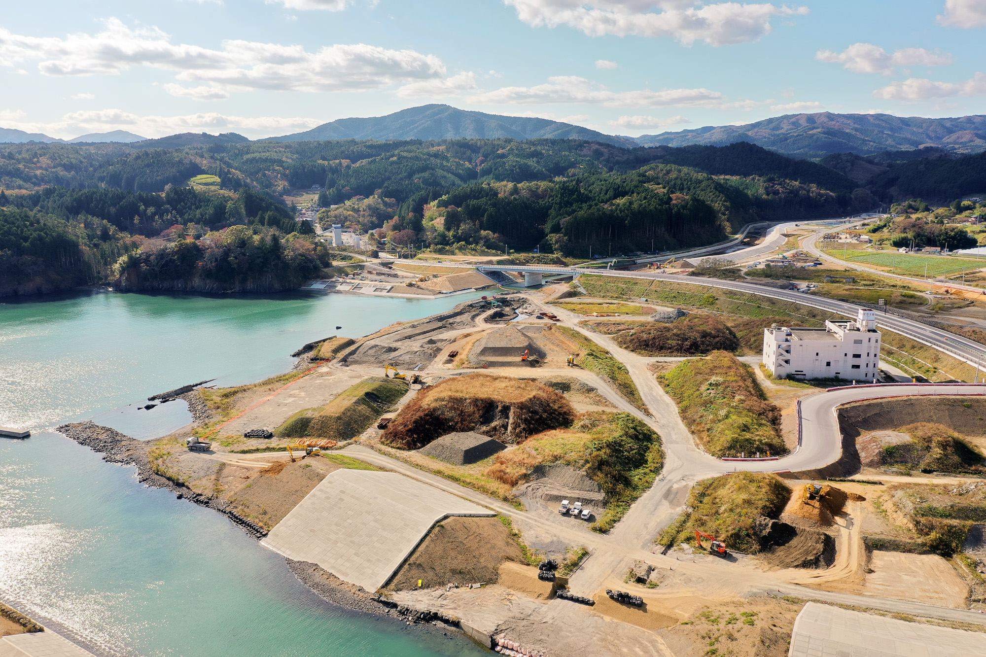 川が海に流れ込む地点で、周囲に工事用重機と盛り土が点在し、背後に山と森林が広がる市の湾奥における復興工事中の写真