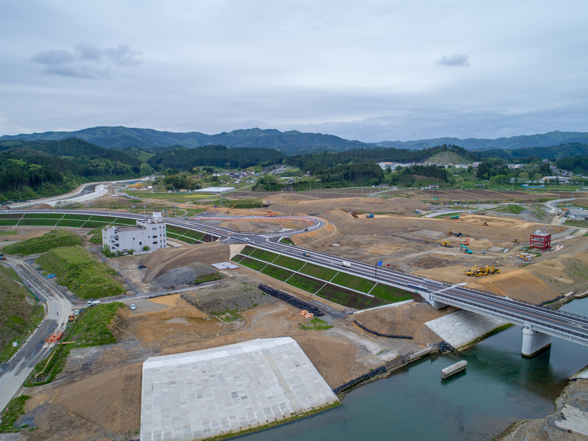 川に架かる橋と、その周辺で道路や通路が整備され、建物や建設機材が点在する工事現場を上空から捉え、背景には緑豊かな丘が広がる風景の写真