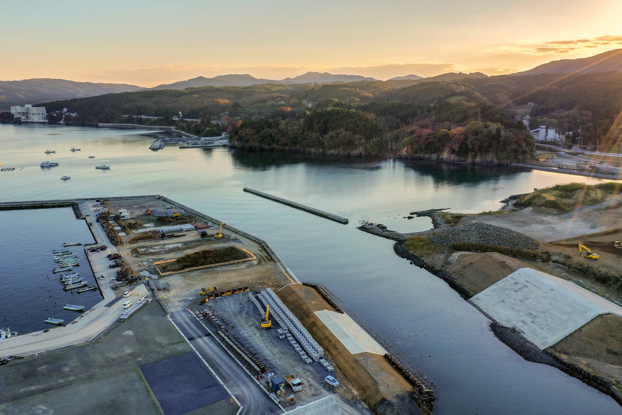 海沿いの風景で、港には複数の船が停泊しており、陸地には工事用の重機が見られ、穏やかな水面が周囲の山や木々を映し出し、空は澄んでいて夕焼けの暖かい光が全体を照らしている写真