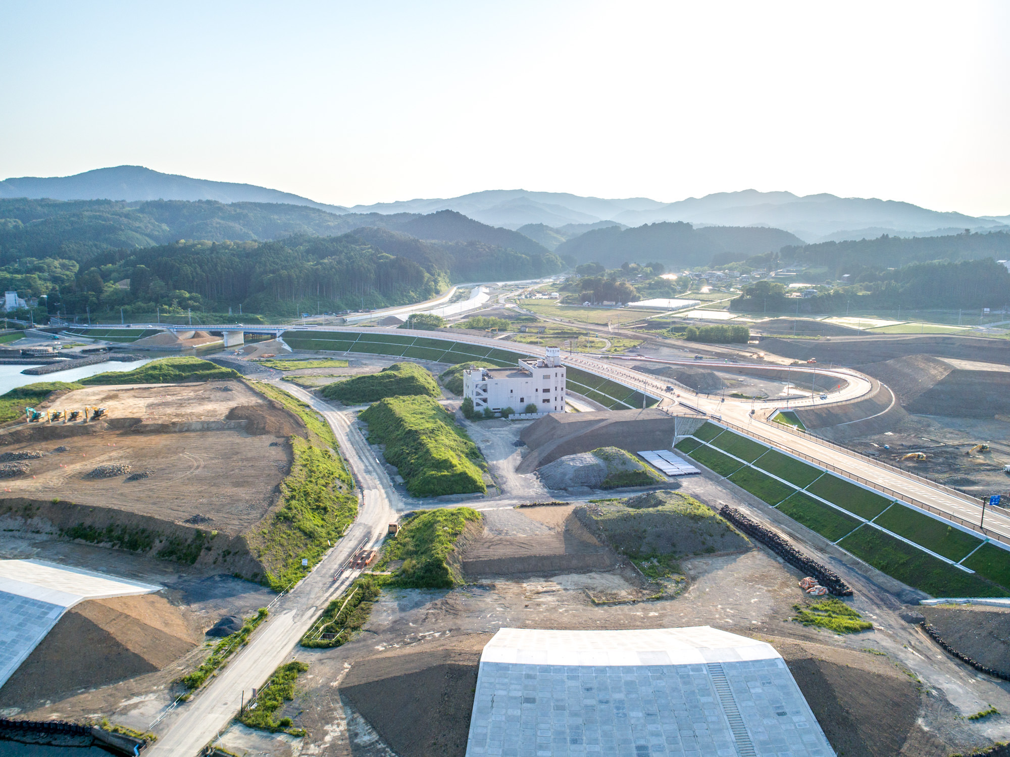 複数の区画が開発中の建設現場を上空から撮影したもので、道路や緑地、土の盛り上がりが点在し、中央には白い建物と建設資材・機材があり、背景にはなだらかな丘と森林、晴れた空が広がる風景の写真