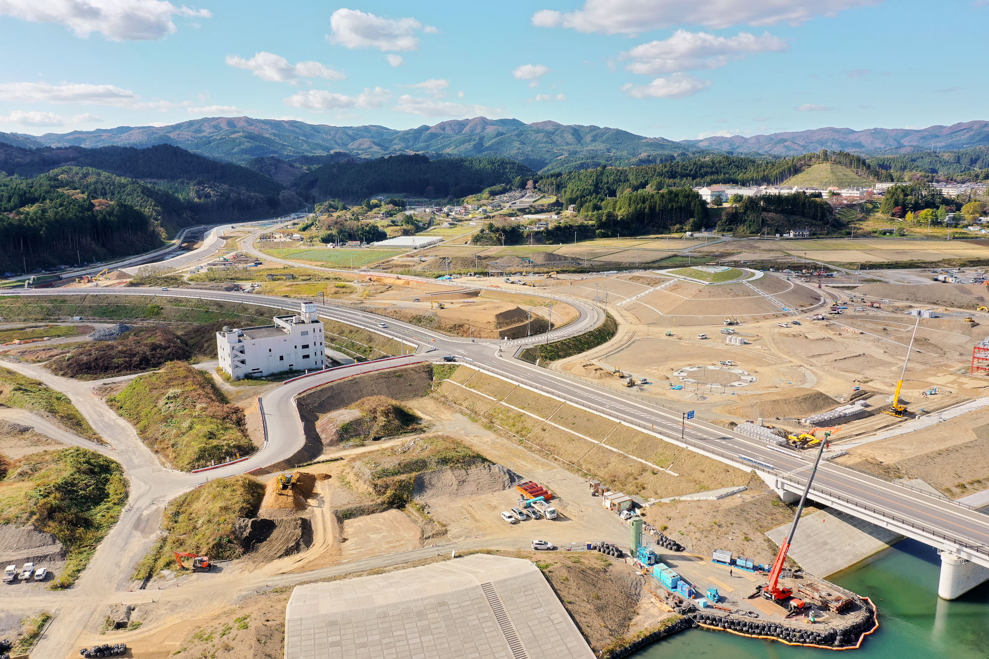 小高い場所にある白い建物とその周囲に造成地や整備中の道路が広がり、背景には里山と遠くの集落が見える復興造成地の写真