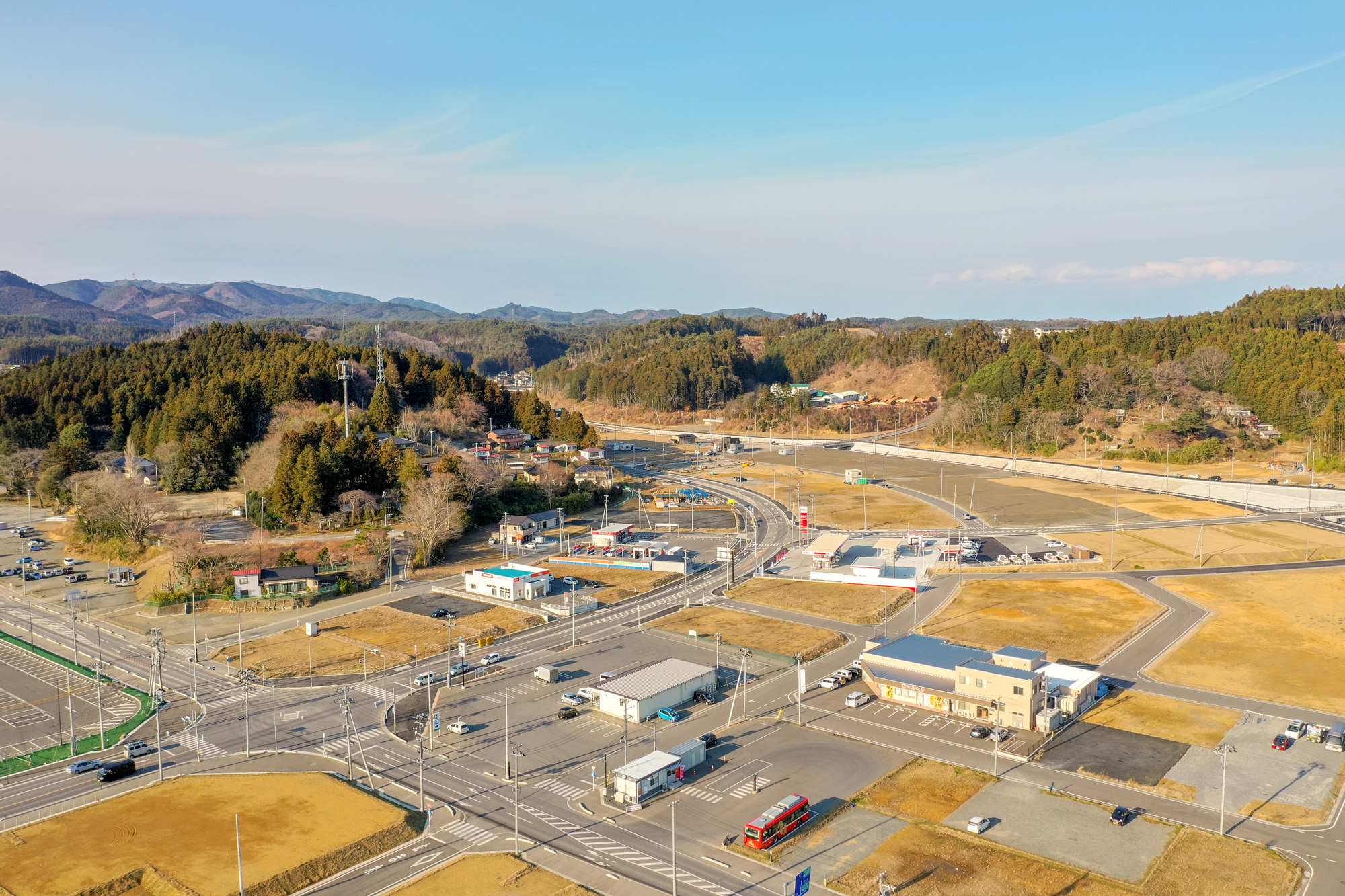 丘や森林に囲まれ中心に建物や道路、交差点が広がり、空き地や駐車場、車両が点在する自然と人の営みが調和した地域を上空から捉えた地方の町の写真